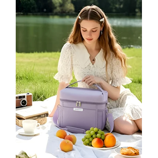 Person holding purple lunch bag with strap on picnic blanket, fruits and cup nearby